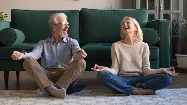 Happy mature couple having fun, practicing yoga together at home, laughing grey haired man and woman sitting in lotus pose on floor in living room, breathing, relaxing, healthy lifestyle concept