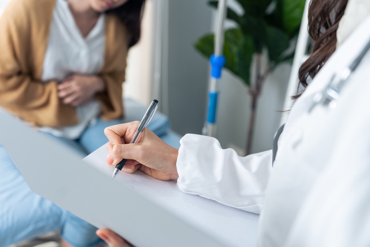 Doctor writing on pad in foreground, with a woman experiencing stomach pain in the background