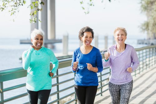 A group of three multi-ethnic women in their 50s and 60s power walking on a city waterfront side by side. They are having a good time, laughing.