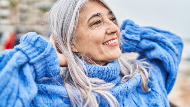 Middle age gray-haired woman smiling with hands on head at seaside, wearing a blue sweater