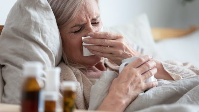 Ill sick middle aged woman sneezing blowing running nose holding tissue sit on bed, upset old mature lady caught cold got flu influenza grippe symptoms drink hot tea taking medications at home alone