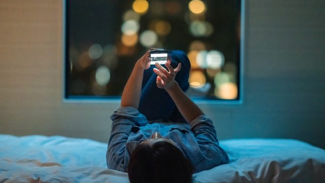 A woman is lying down on a bed and using a smart phone at night.