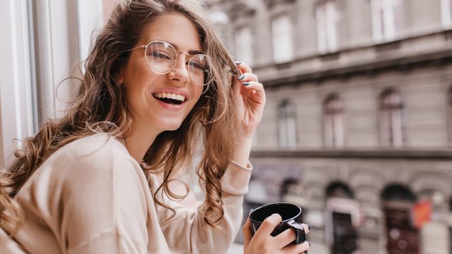 Close-up portrait of laughing brunette girl in beige sweater drinking coffee on city background.