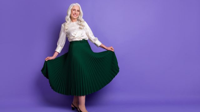 A happy mature woman with long white hair wearing a white silk blouse and long emerald green skirt against a purple background