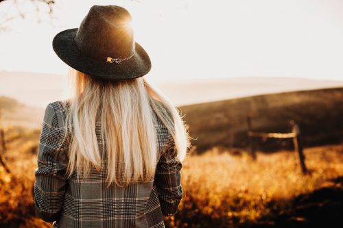 Back view of a lovely blonde girl dressed jacket and hat and looking at epic view at the sunset.