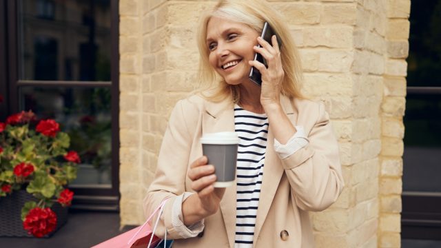 Stylish older woman smiling on phone with trench coat, holding a shopping bag and coffee