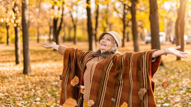 Happy senior woman wearing a poncho enjoying an autumn day in the park and throwing leaves.