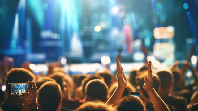 Rear view of large group of people enjoying a concert performance. There are many hands applauding and taping the show.