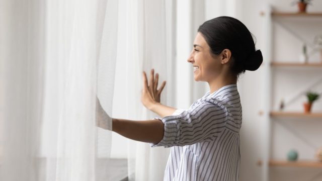A young brunette woman opens her sheer window curtains and smiles.