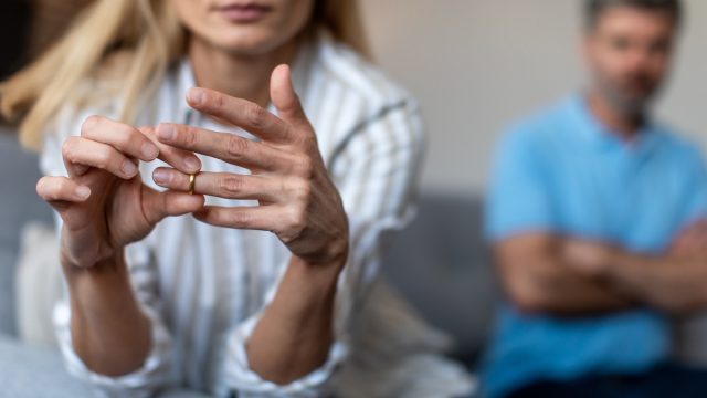 Close up of a woman removing her wedding band, with a crossed-arm man in the background.
