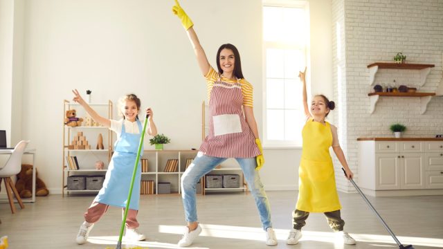 Happy family having fun while cleaning their house. Cute little children helping mom tidy up their apartment. Smiling young mother and daughters dancing together while mopping the floor at home