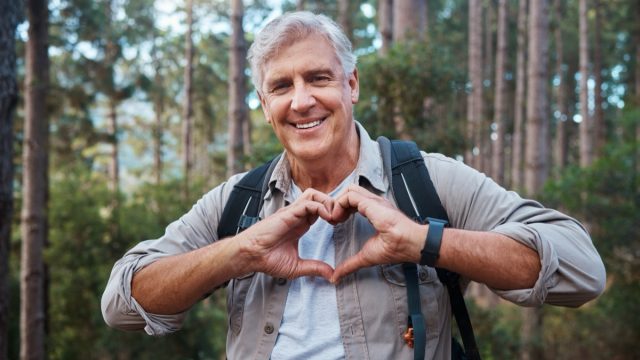 Portrait, heart and senior man in forest hiking, fitness and health support, self care hands and cardiovascular workout. Nature, travel or trekking of happy person in woods with love sign or emoji