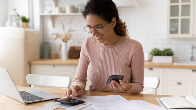 Young female work with financial papers at home count on calculator before paying taxes receipts online by phone. Millennial woman planning budget glad to find chance for economy saving money