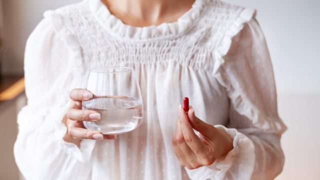 Woman in white blouse holding in hand iron Ferrum supplement capsule and glass of water. Bioactive additive woman pharmacy. Vitamin mineral treatment against anemia. Autumn health care concept