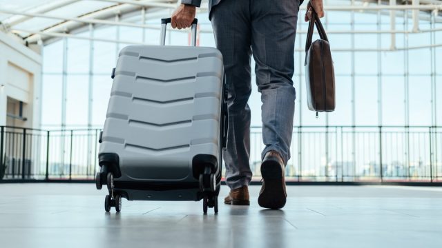 Low angle view of unrecognizable businessman in gray suit walking through airport terminal with briefcase and suitcase.