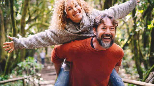 Overjoyed adult couple have fun together at outdoor park in leisure activity. Man carrying woman in piggyback and laugh a lot. Love and life mature people lifestyle concept. Enjoying vacation nature