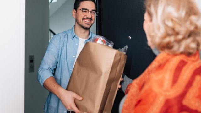 A young male volunteer handing a grocery bag at the door to an older woman