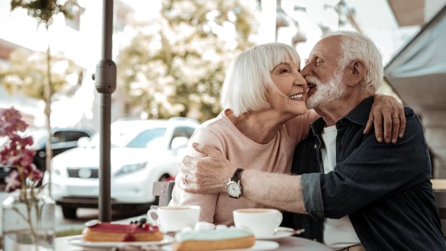 A happy senior couple kissing while sitting at an outdoor cafe with cake on the table