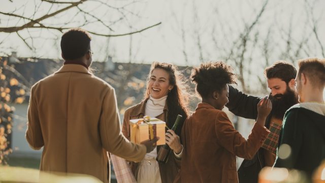 Candid shot of cheerful young couple welcoming and greeting their friends who came to their home as guests. They are standing outside, in warm autumnal sunshine.