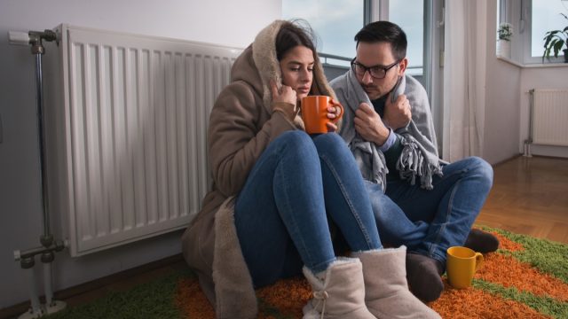 Young couple in jacket and covered with blanket sitting on floor beside radiator and trying to warm up