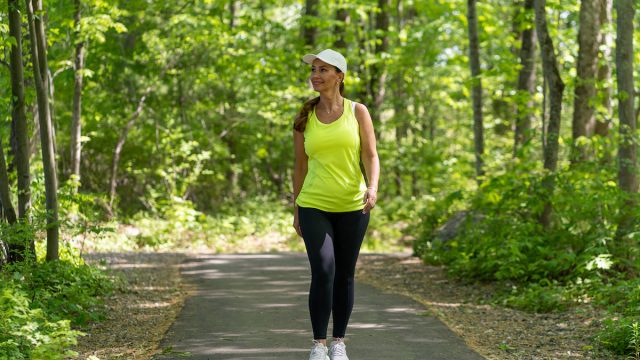 A middle-aged woman wearing black leggings and a bright yellow tank is taking a walk along a tree-lined path