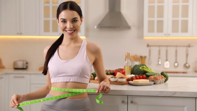 Happy woman measuring waist with tape in kitchen