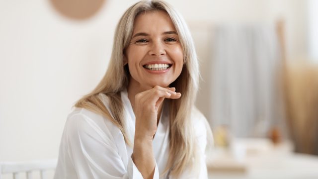 Happy attractive blonde woman in white silky bathrobe posing at white bathroom, smiling at camera, closeup portrait, panorama with copy space. Face and skin care at home concept