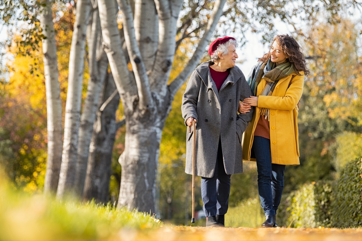 young woman wearing a yellow coat holding the arm of a senior woman wearing a gray coat and using a cane while they take a walk outside on a fall day