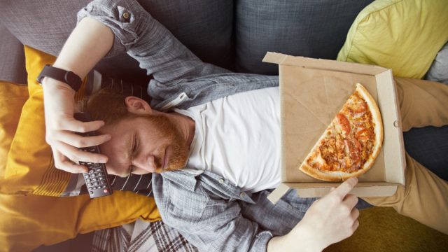 Above view portrait of bearded man lying on couch and eating pizza while watching TV at home, copy space