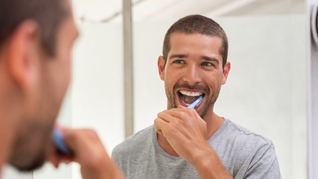 Smiling young man with toothbrush cleaning teeth and looking mirror in the bathroom. Handsome man brushing his teeth in morning in bathroom. Happy guy in pajamas brushing teeth before going to sleep.