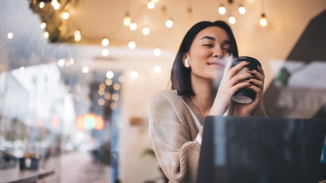 Asian girl drinking and enjoying coffee during working on laptop in cafe. Young millennial brunette woman wearing earphones sitting at table. Freelance and remote work. Modern female lifestyle
