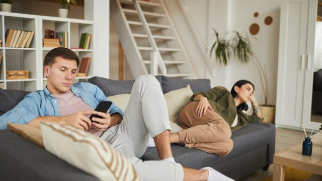 bored young couple watching TV on sofa at home, focus on man using smartphone in foreground
