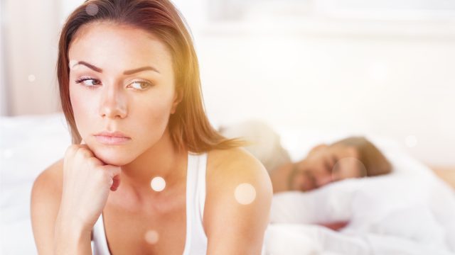 A woman sits on a bed next to a sleeping man.