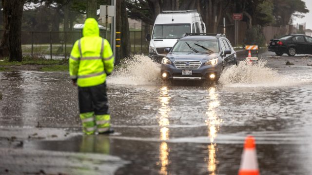 A traffic guard in hi-vis gear guides a car away from a flooded area in a street