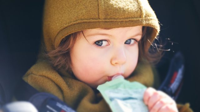 Close up portrait of sweet toddler kid eating fruit puree from plastic doy pack, sitting in stroller, outdoor snack time