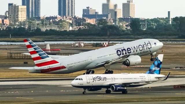 A JetBlue plane taxiing on a runway while an American Airlines jumbo jet takes off in the background