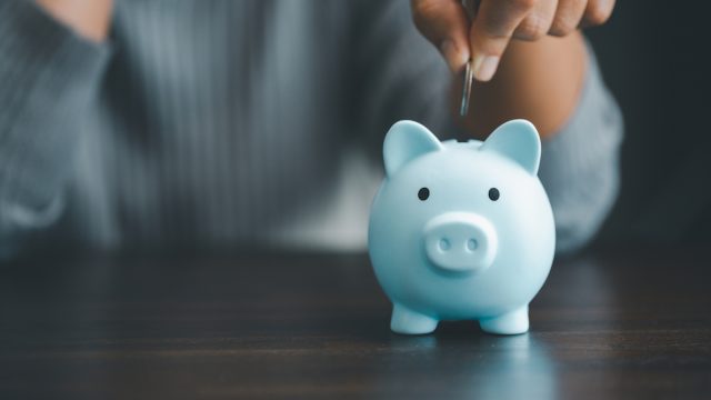 Blurred image of a person wearing a blue sweater putting a coin into a blue piggy bank.