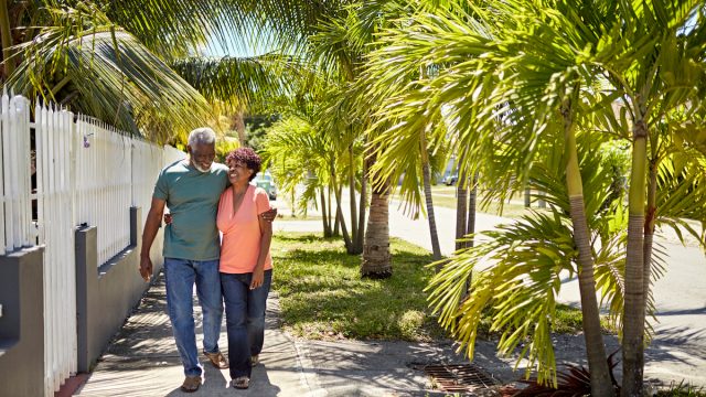 A senior couple hugging and walking down a sidewalk lined with palm trees on a sunny day.