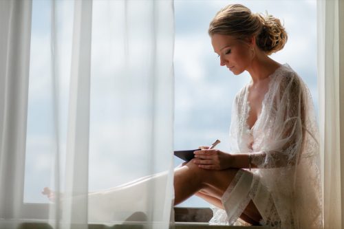 Bride in lace robe sitting in window and writing in a notebook