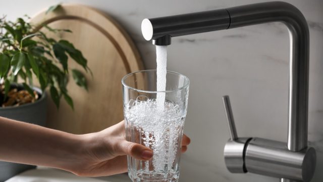 Woman filling glass with tap water from faucet in kitchen, closeup
