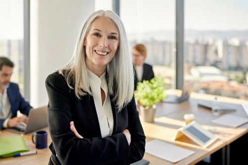 Waist-up front view of longhaired executive in businesswear sitting on table with arms crossed and smiling at camera as colleagues work in background.