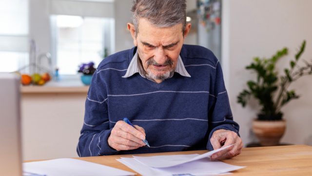 A senior adult man sitting at a table stacked with papers. He signs the documents and pays the bills