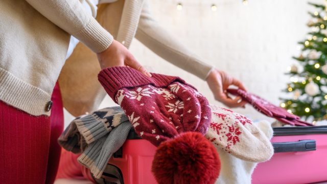 Christmas time. Woman putting knitted winter clothing in a suitcase in the room decorated christmas tree. Travel, holiday