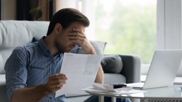 Upset man with his head in his hand holding a form while sitting on the floor in front of his laptop.