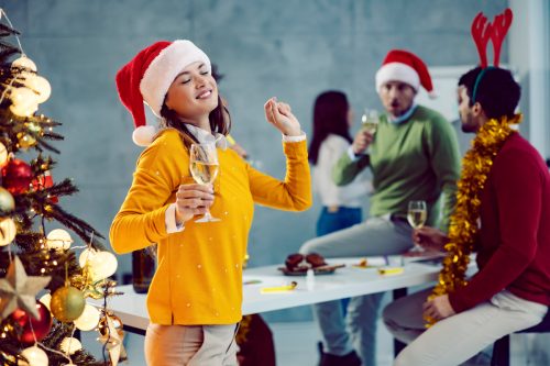 A young businesswoman is standing next to a Christmas tree in the boardroom with a glass of champagne in her hands and dancing alone.