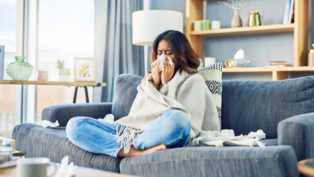 A woman sitting on the couch wrapped in a blanket and blowing her nose while surrounded by used tissues, possibly sick with the flu or COVID