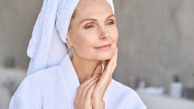 Happy beautiful middle aged woman wearing bathrobe and white towel with perfect complexion touching face looking away in bathroom. Advertising of skin care spa wellness concept. Closeup portrait.