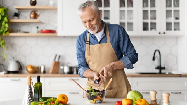 Happy senior man preparing healthy vegetable salad in kitchen, seasoning meal, smiling elderly gentleman in apron adding salt to bowl, enjoying cooking vegetarian food at home, free space