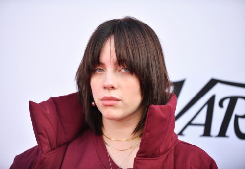 Billie Eilish posing at a Variety event wearing a red coat with a large collar; her brunette hair is styled in a wolf cut