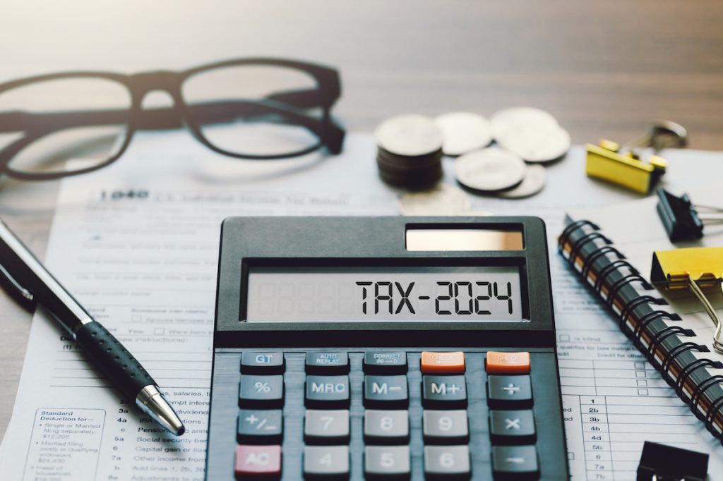 "Tax 2024" written out on a calculator among paperwork, glasses, pen, and coins.
